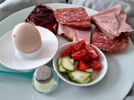 Frühstück im Bett mit gekochtem Ei,Salami-, Schinken- und Marmeladebroten sowie Gurke und kleine Toatenvierten